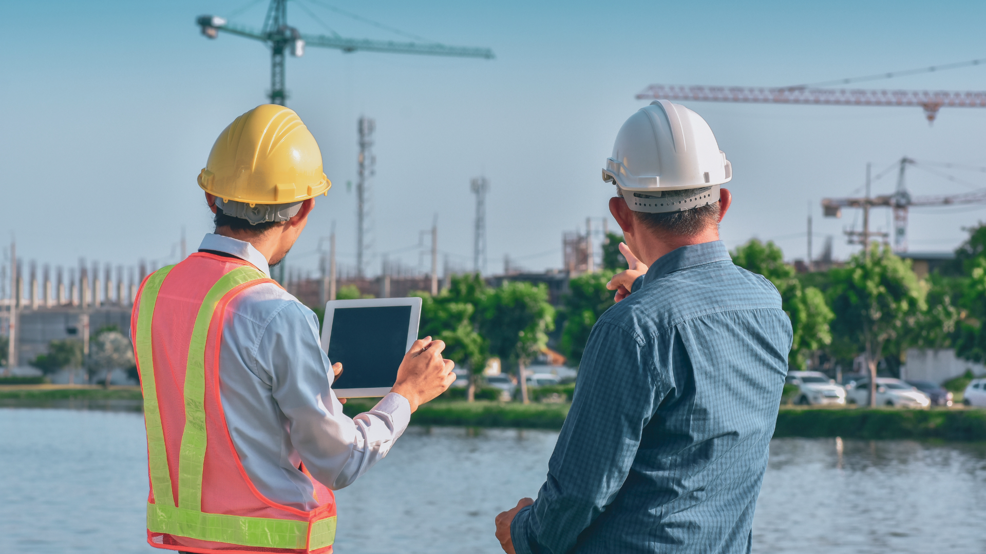 Zwei Bauleiter, einer davon mit einem Tablet in der Hand, stehen vor einer Baustelle. Im Hintergrund erkennt man Kräne und Baugerüste.